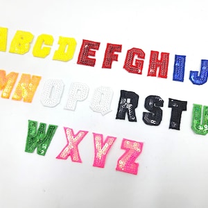 以下が含まれることがあります： さまざまな色のスパンコールでできたアルファベット26文字のセット。文字はAからZまで、行に沿って配置されています。文字はすべて大文字で、高さは約5cmです。