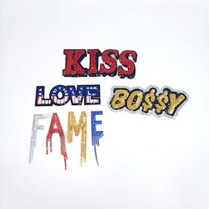 Op de afbeelding: Een set van vijf strijkbare patches met pailletten-lettering. De patches spellen "KISS", "LOVE", "FAME" en "BOSSY". De patches zijn in rood, wit, blauw, goud en zilver.