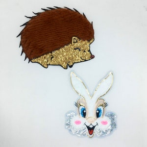 Könnte beinhalten: Zwei dekorative Aufnäher: ein brauner Igel mit goldener Paillettenbauch und ein weißes Hasengesicht mit blauen Augen, rosa Wangen und silberner Paillettenborte. Diese Aufnäher sind wahrscheinlich für Kleidung oder Bastelprojekte gedacht.