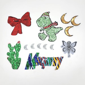May include: A set of iron-on patches featuring a green cactus, a red bow, a green dinosaur wearing a red and white scarf and a Santa hat, a white fairy with black wings, a silver crescent moon, and a multi-colored sequin word "Merry".