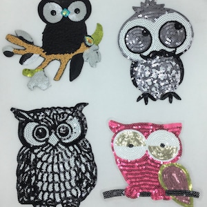 May include: Four sequined owl appliques in different colors and designs. The owls are black, gray, pink, and black and white. They are all wearing a branch.