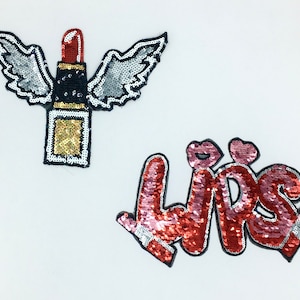 May include: Two iron-on patches with a sparkly sequin design. One patch features a red lipstick with gold and silver wings. The other patch says "Lips" in pink and red sequins.