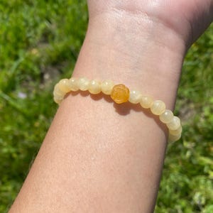 May include: A yellow beaded bracelet with a rose-shaped charm. The bracelet is made of small, round beads and has a single, larger bead in the center that is shaped like a rose.