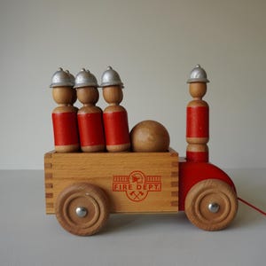 Puede incluir: Juguete de madera para tirar con diseño de camión de bomberos. El juguete tiene una caja de madera con la inscripción "FIRE DEPT.", cuatro figuras de madera con cascos plateados y uniformes rojos, y ruedas de madera. Una cuerda roja está unida.
