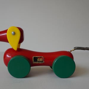 Puede incluir: Un juguete de perro de madera roja para tirar, con ruedas verdes y una oreja amarilla. El perro tiene una cola de resorte con una bola amarilla al final. El juguete tiene las letras "BR" en el lateral.