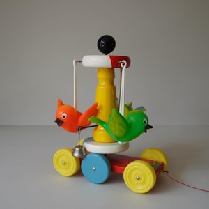 May include: A colorful wooden pull-along toy featuring a red base with yellow wheels, a blue wheel, and a central yellow column. Two bird figures, one orange and one green, are suspended from the top. A small silver bell hangs below the orange bird.
