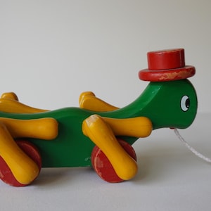 Könnte beinhalten: Ein grünes Holz-Zugspielzeug in Form einer Schildkröte mit gelben Beinen und roten Rädern. Es hat einen roten Zylinderhut und eine weiße Schnur. Das Spielzeug ist für kleine Kinder konzipiert.