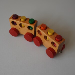 Puede incluir: Dos trenes de juguete de madera con ruedas rojas y clavijas de colores. Cada vagón tiene un cuerpo rectangular con agujeros circulares y clavijas en tonos rojos, amarillos, verdes y naranjas. Los trenes son de madera clara.