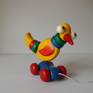 May include: A colorful wooden pull-along toy bird. The bird is primarily yellow with red, green, and blue accents. It has large white eyes, a red beak, and a red and orange wing. The bird sits on a blue base with red wheels and a white pull string.
