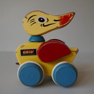 May include: A vintage wooden pull-along duck toy. The duck is primarily yellow with a red beak and wings. It has blue wheels and a blue neck. The toy has the brand name "BRIO" on the side and is marked "MADE IN SWEDEN".