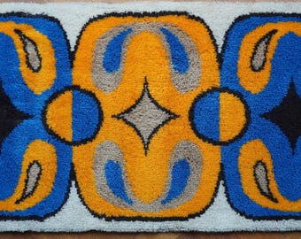 May include: A rectangular rug with a repeating pattern of blue, yellow, and gray shapes. The design includes stylized floral or abstract motifs, with a black diamond shape in the center of each blue section. The rug has a white border.
