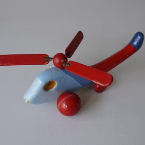 May include: A vintage wooden toy helicopter painted in red, blue, and yellow. The helicopter has a red body, red propeller blades, and a blue and yellow cockpit. The tail is red with a blue tip.