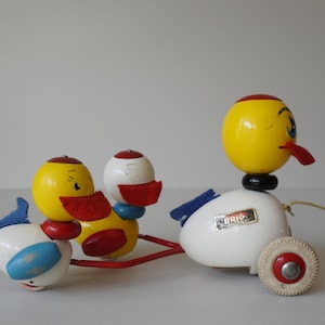May include: A vintage wooden pull-along toy duck family. The ducks have yellow, red, white, and blue painted details. The lead duck has a yellow head, a red beak, and a black neck. The toy has a red metal frame and white wheels.