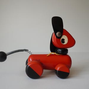 Può includere: Un giocattolo vintage a forma di cane da trascinare, in legno. Il cane è principalmente rosso con dettagli neri sulle orecchie, sul collare e sulle ruote. Ha una coda a molla e una palla nera alla fine.