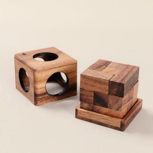 May include: Two wooden puzzles. One is a cube with three circular holes. The other is a cube puzzle made of several wooden blocks. Both are made of a dark brown wood with visible grain.