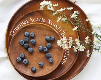 May include: Three round acacia wood plates of varying sizes, stacked on top of each other. The plates are a warm brown color and have a natural wood grain. Blueberries are scattered on the middle plate. The text "Gourmet Acacia Wood Char Plates" is visible.