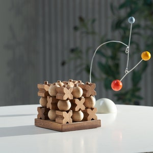 May include: A wooden tic-tac-toe game with a cube design. The game features wooden X and O pieces and a dark brown base. A colorful mobile with red, yellow, and blue spheres hangs in the background. The game is on a white surface.