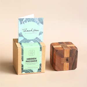 May include: A wooden puzzle cube and a cardboard box with the text "HIDDEN PASSAGE" and a "Thank You" card with a blue leaf design. The puzzle is made of various wood tones.