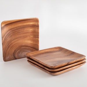 May include: A stack of three square wooden plates with rounded corners, next to a single plate. The plates are a warm brown color with a visible wood grain pattern. The plates are likely for serving food.