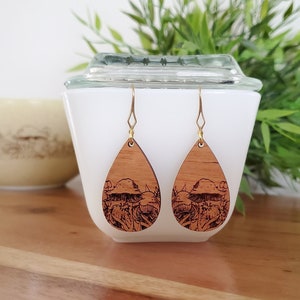 May include: Teardrop-shaped wooden earrings featuring a laser-etched mushroom design. The earrings are light brown and suspended from gold-coloured ear wires. They are displayed in front of a white container, with a green plant visible.