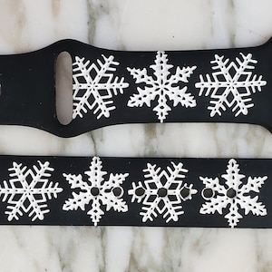 May include: Black silicone Apple Watch band with white snowflake designs. The band is shown in two pieces, one with a silver buckle and the other with a silver clasp.