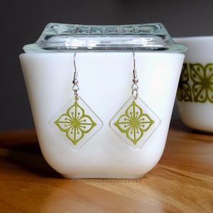 Puede incluir: Un par de pendientes con forma de diamante con un diseño de flor en el centro. Los pendientes son de color verde y blanco con ganchos plateados.