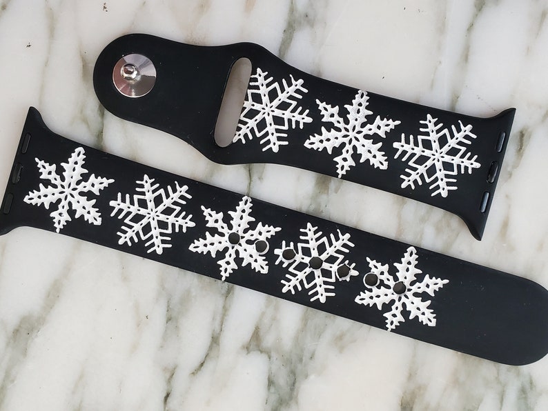 May include: Two black silicone watch bands with white snowflake designs. The bands are for Apple Watches and have a classic buckle closure.