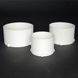 Może przedstawiać: Trzy białe plastikowe pierścienie do dekorowania tortów w różnych rozmiarach. Pierścienie mają gładką, zaokrągloną powierzchnię i lekko fakturowane dno.