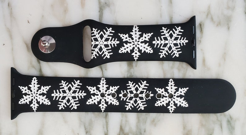 May include: Black silicone Apple Watch band with white snowflake designs. The band is shown in two pieces, one with a silver buckle and the other with a silver clasp.