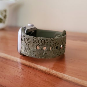 May include: A green silicone watch band with a floral pattern. The band is designed for a smartwatch and has a silver buckle.