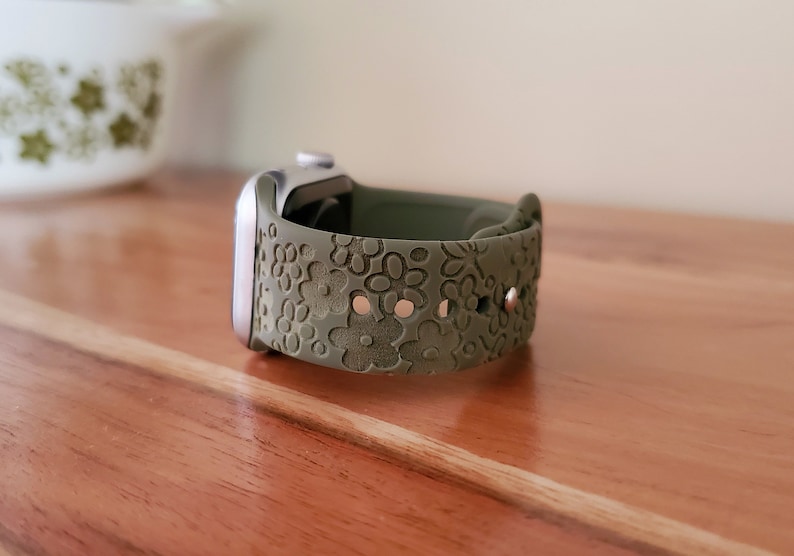 May include: A green silicone watch band with a floral pattern. The band is designed for a smartwatch and has a silver buckle.