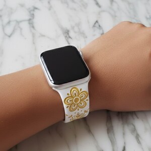 An AI-generated lifestyle mockup showing a white silicone Apple Watch band with a Pyrex Buttefly Gold  floral pattern on a model's wrist, displayed over a white marble background to show scale and fit.