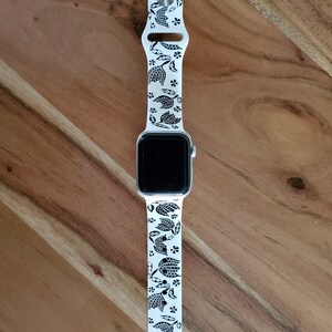 Może przedstawiać: Biały zegarek Apple Watch z czarno-białym paskiem w kwiatowy wz&oacute;r. Tarcza zegarka jest czarna, a pasek wykonany jest z silikonu.