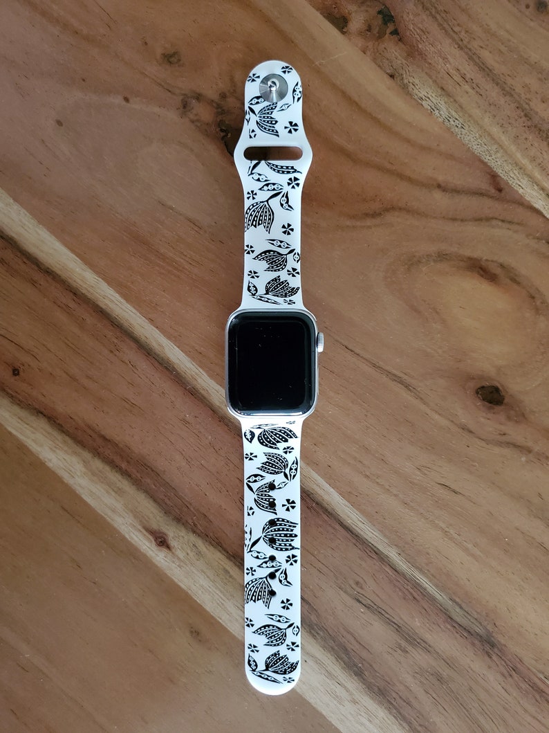 Może przedstawiać: Biały zegarek Apple Watch z czarno-białym paskiem w kwiatowy wz&oacute;r. Tarcza zegarka jest czarna, a pasek wykonany jest z silikonu.