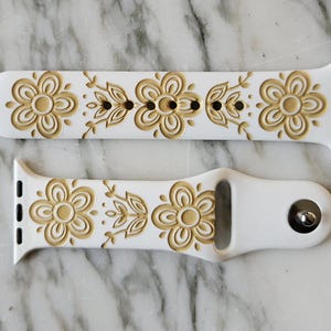 White silicone Apple Watch band with a floral design. The bands feature the Pyrex Butterfly Gold pattern of embossed flowers and leaves in a goldenrod color. The bands are shown on a marble surface.