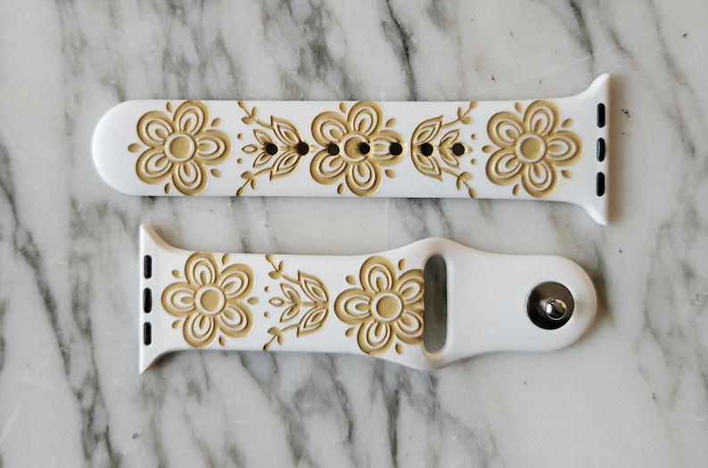 White silicone Apple Watch band with a floral design. The bands feature the Pyrex Butterfly Gold pattern of embossed flowers and leaves in a goldenrod color. The bands are shown on a marble surface.