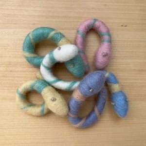 May include: Five felt snake toys in different colors and patterns. The snakes are curled up together on a wooden surface. The colors are blue, green, yellow, and pink.