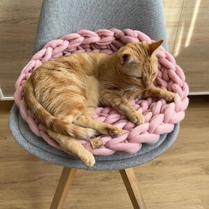 May include: An orange tabby cat resting in a pink, chunky knit cat bed on a gray chair. The cat bed is round and has a woven texture. The chair has wooden legs.