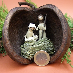 Può includere: Una scena della natività realizzata con figure di paglia e stoffa all'interno di una mela scavata. Le figure sono una donna, un uomo e un bambino. La mela è marrone e ha una consistenza ruvida.