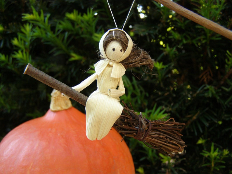 May include: A handmade witch doll made of natural materials, sitting on a twig broom. The doll has a white dress and a brown hair. The background includes a large orange pumpkin and green foliage, suggesting a fall or Halloween theme.