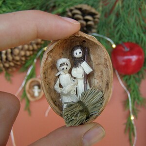 Può includere: Una scena della natività in miniatura realizzata con materiali naturali, tra cui un guscio di noce, paglia e stoffa. Le figure sono quelle di Maria, Giuseppe e il bambino Gesù, e sono disposte all'interno del guscio di noce.