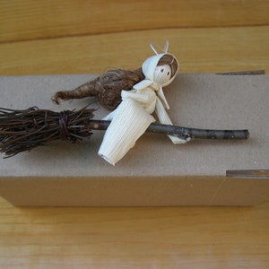 May include: A handmade miniature witch figurine riding a broom. The witch is crafted from natural materials, including straw and corn husks, and is perched on a twig broom. The broom is made of twigs bound together with twine. The witch is on a cardboard box.