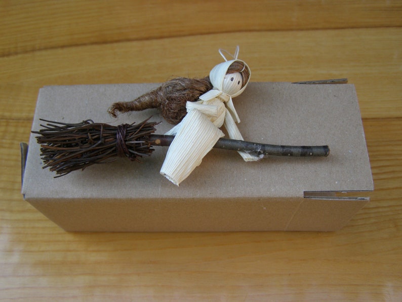 May include: A handmade miniature witch figurine riding a broom. The witch is crafted from natural materials, including straw and corn husks, and is perched on a twig broom. The broom is made of twigs bound together with twine. The witch is on a cardboard box.
