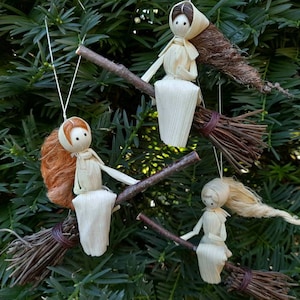 May include: Handmade witch ornaments crafted from natural materials. Each witch doll sits on a twig broom, with straw hair and a simple dress. The ornaments are suspended against a backdrop of green foliage.