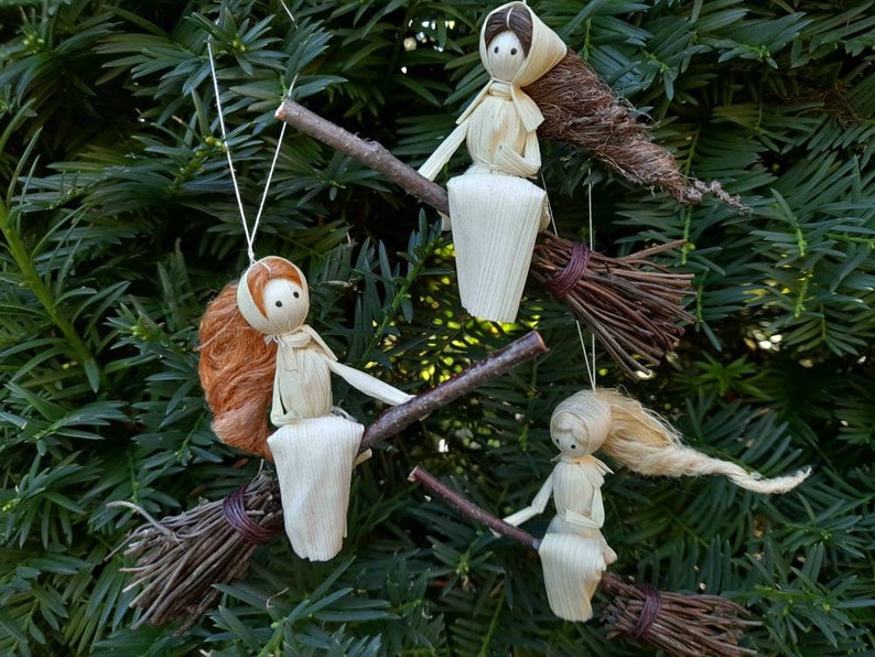 May include: Handmade witch ornaments crafted from natural materials. Each witch doll sits on a twig broom, with straw hair and a simple dress. The ornaments are suspended against a backdrop of green foliage.