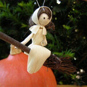 May include: A handmade witch doll made from corn husks, riding a twig broom. The doll has a white dress and a brown hair and hat. The background is a blurred green foliage and a large orange pumpkin.