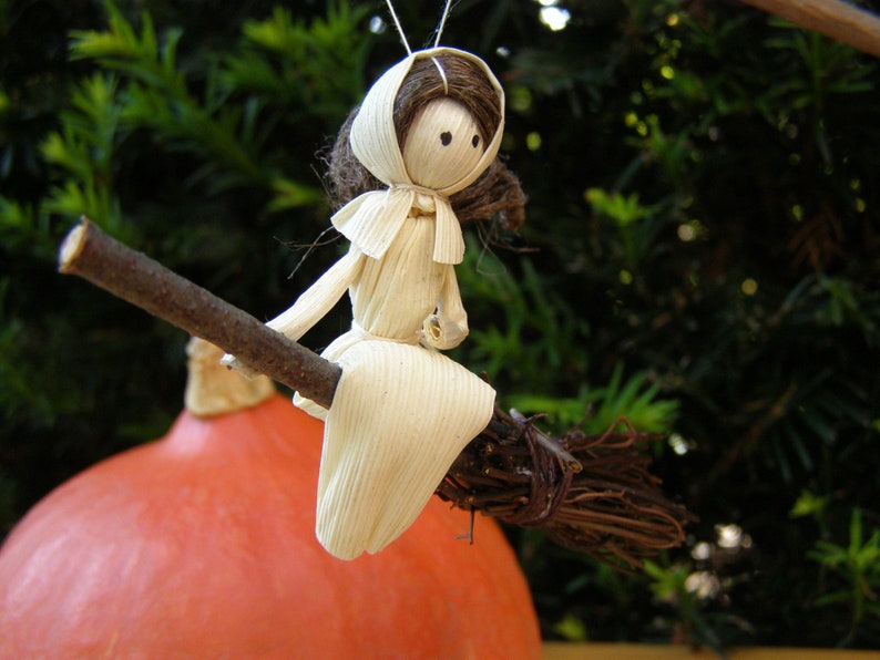 May include: A handmade witch doll made from corn husks, riding a twig broom. The doll has a white dress and a brown hair and hat. The background is a blurred green foliage and a large orange pumpkin.