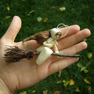 May include: A handmade miniature witch riding a broom. The witch is crafted from natural materials, including straw and paper, with a brown broom and a white hat. The background is a blurred green lawn.