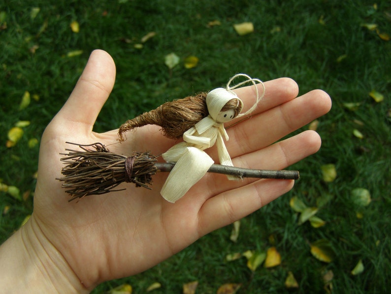 May include: A handmade miniature witch riding a broom. The witch is crafted from natural materials, including straw and paper, with a brown broom and a white hat. The background is a blurred green lawn.