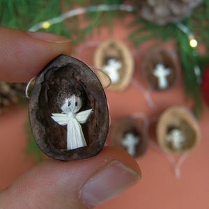 Può includere: Ornamenti natalizi fatti a mano con minuscole figure di angeli realizzate in materiale bianco, annidate all'interno di gusci di noce. Gli angeli hanno ali e sono esposti su uno sfondo sfocato con decorazioni festive.